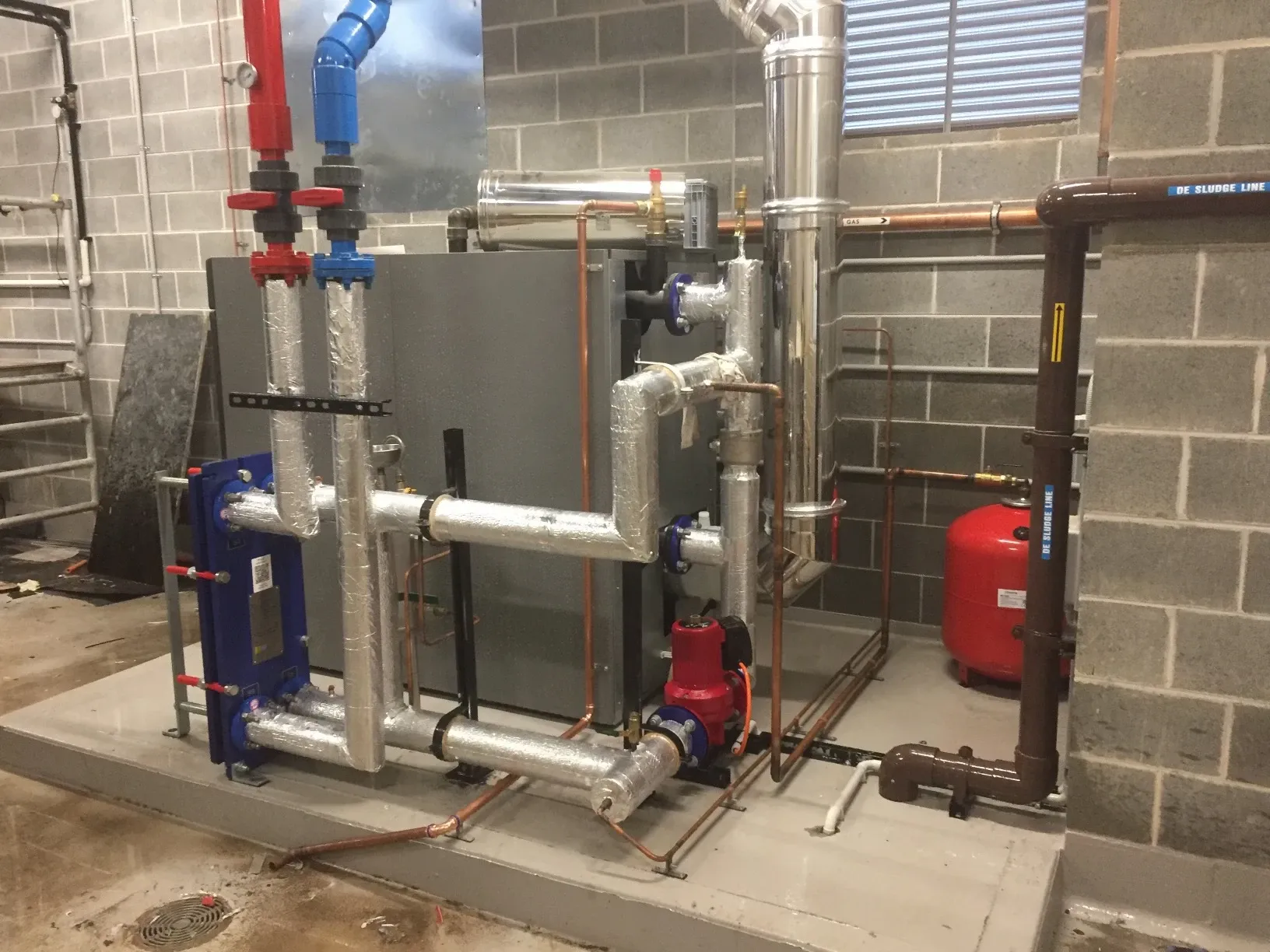 Commercial hydronic plant with a blue plate heat exchanger, red circulating pumps and insulated flow and return pipework in a plant room
