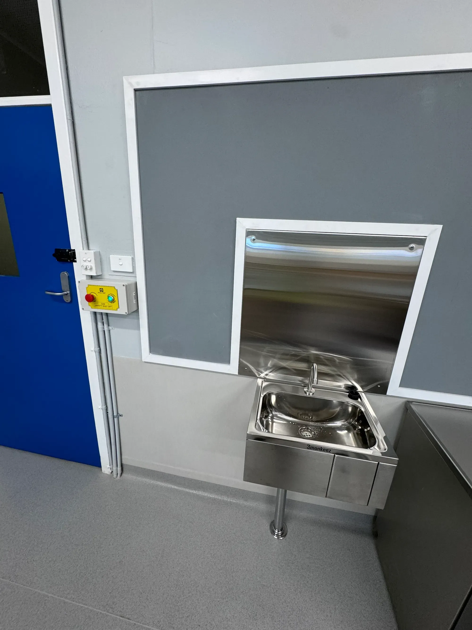 Yellow gas safety shut-off panel with emergency stop beside a wall-hung knee-operated stainless hand basin in a commercial food-prep room